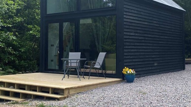 A modern, black wooden cabin with large glass windows, a wooden deck, and a seating area with chairs.