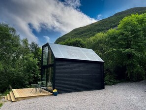 Una cabaña moderna de madera oscura con un añadido de cristal, rodeada de grava y vegetación.