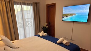 Een hotelkamer met een bed, een televisie waarop een uitzicht op het strand te zien is, een houten deur en een raam met gordijnen.
