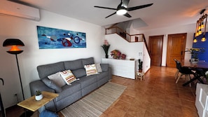 Ein Wohnzimmer mit grauem Sofa, einem Gemälde, einem Deckenventilator, einer Treppe und einem Essbereich.