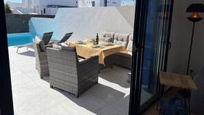 Een terras met rieten meubilair, een tafel gedekt voor twee personen en uitzicht op een zwembad en gebouwen op de achtergrond.