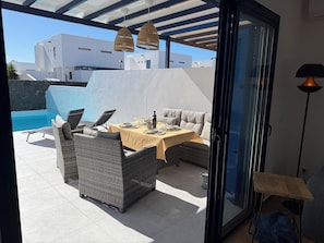Ein Essbereich im Freien mit Korbmöbeln, einem für zwei Personen gedeckten Tisch und Blick auf einen Pool sowie Gebäude im Hintergrund.