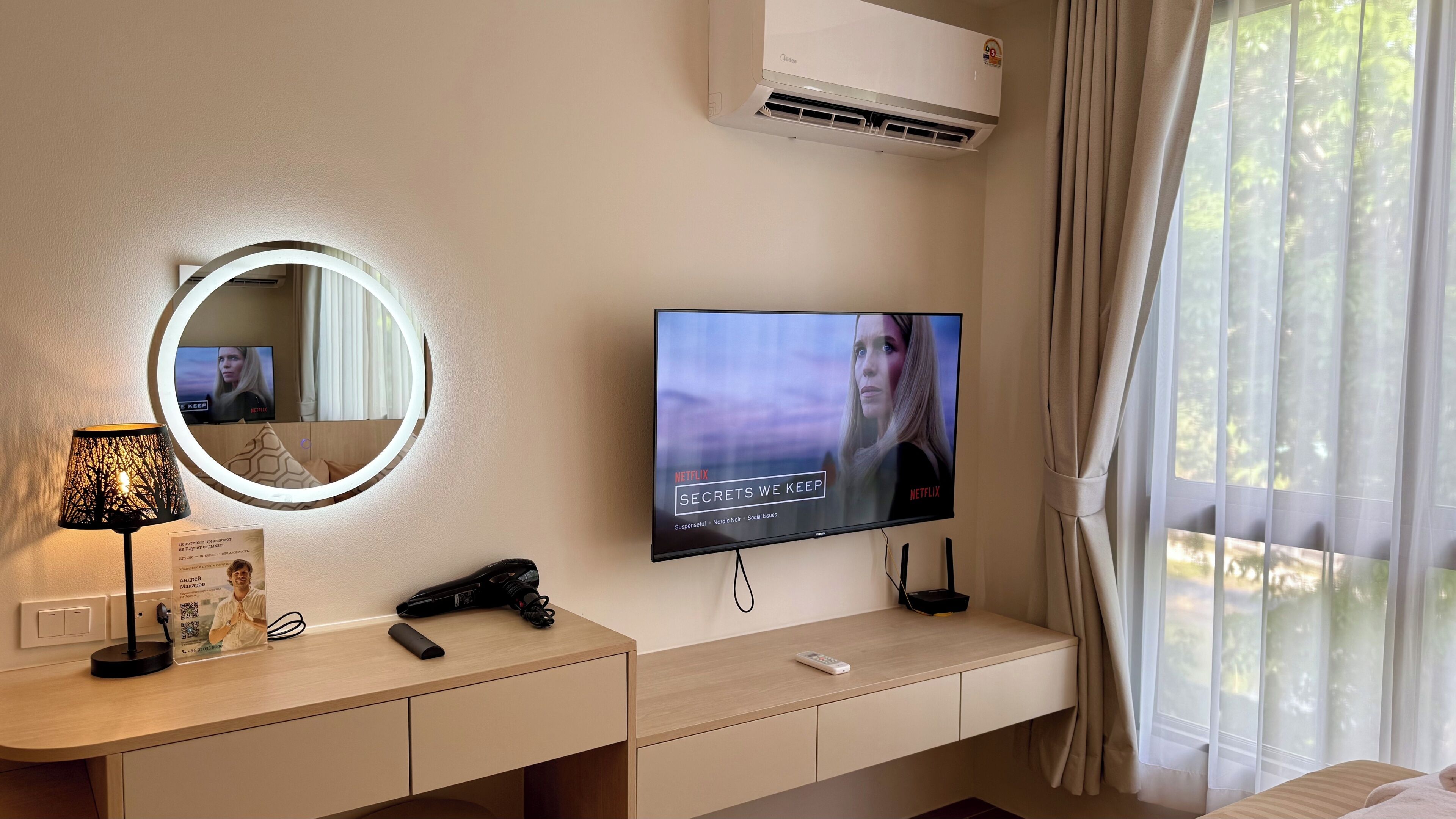 Quarto de hotel com televisão exibindo um programa da Netflix, luminária de cabeceira, secador de cabelo, controle remoto e unidade de ar condicionado.