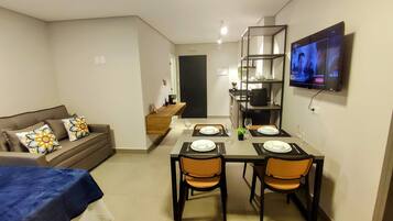 Un espacio habitable compacto con comedor, un sofá con cojines estampados, un televisor montado en la pared y una cocina al fondo.