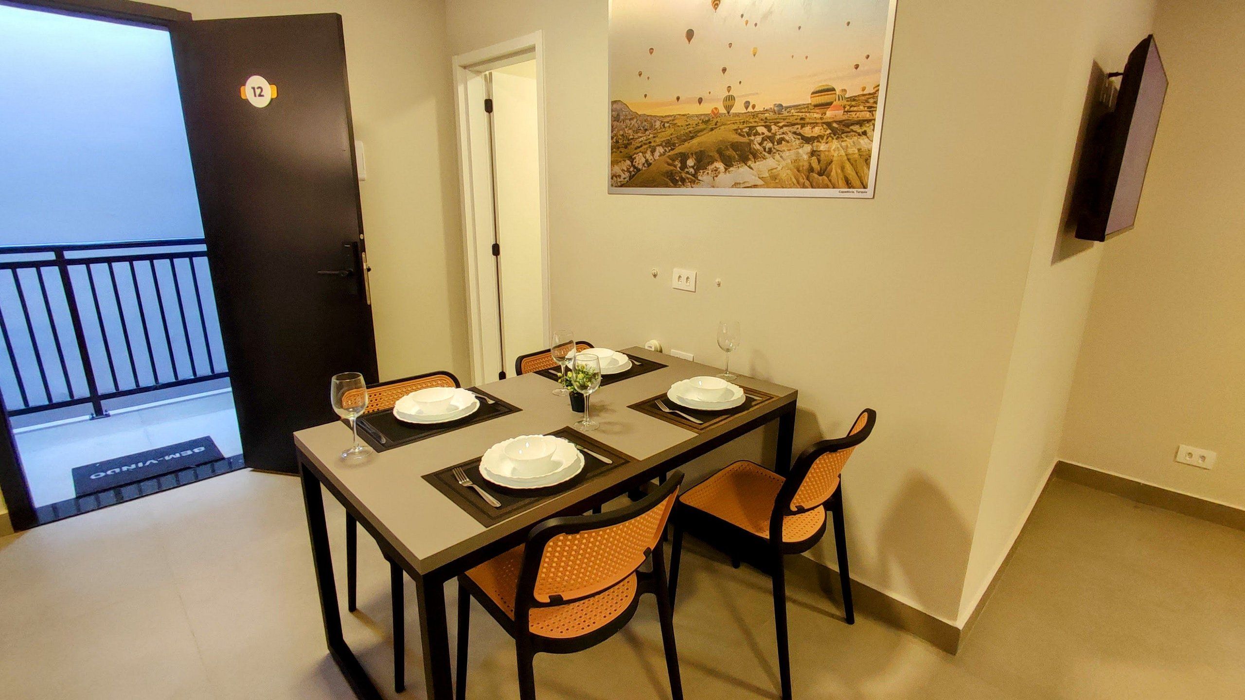 A dining area with a table set for two, two chairs, a door with a number, and a wall-mounted TV.