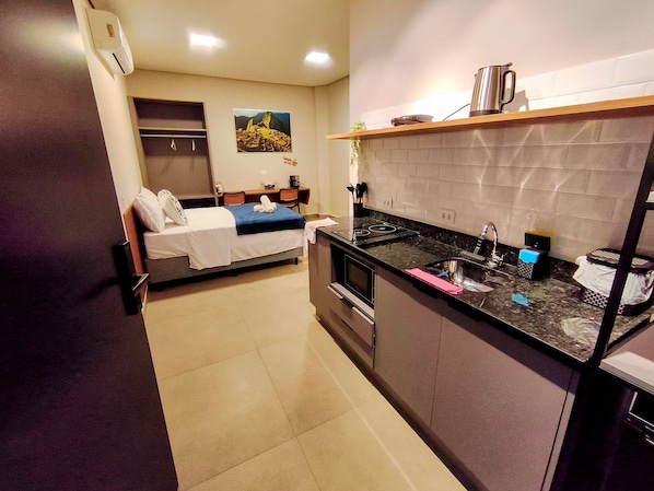 A compact hotel room with a bed, a small desk, a kitchenette, and a bathroom.