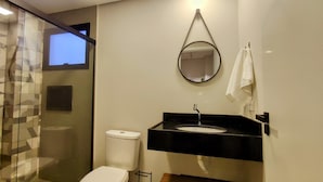 A bathroom with a glass shower enclosure, a toilet, a sink with a black countertop, and a mirror with a towel hanging on the wall. - Airport Residence (Guarulhos)