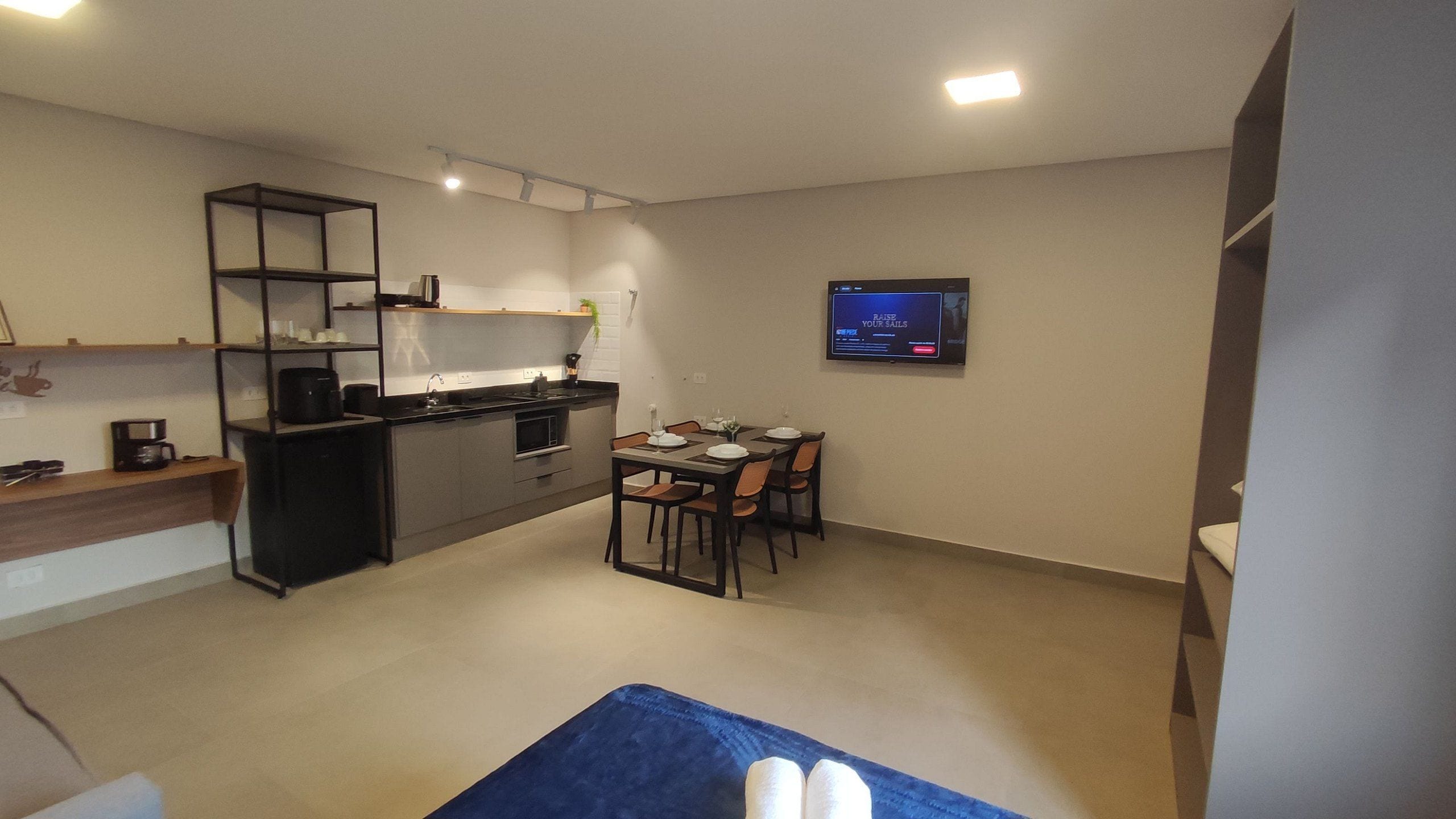 A compact hotel room with a kitchenette, a dining area, and a flat-screen TV mounted on the wall.