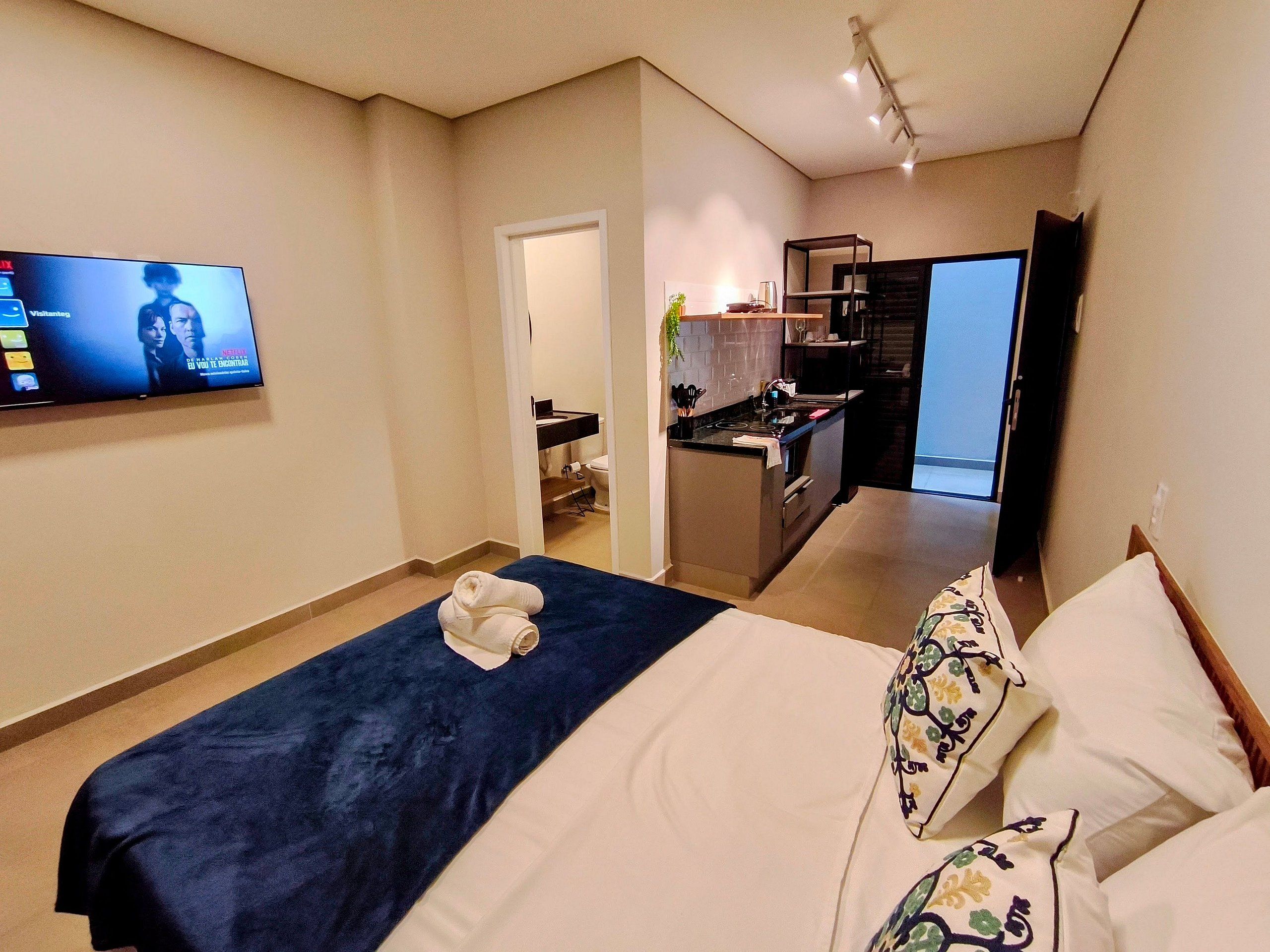 Una habitación de hotel con una cama, un televisor montado en la pared, una kitchenette y un baño visible a través de una puerta abierta.