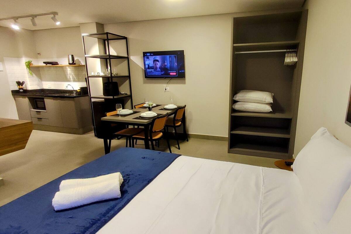 Quarto de hotel com cama, mesa de jantar com cadeiras, televisão, armário e um quadro na parede.