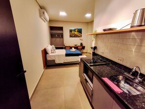 A compact hotel room with a kitchenette, a bed with a blue blanket, a dining area, and a painting on the wall.
