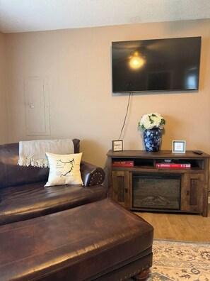 Ein Wohnzimmer mit einer Ledersofa, einem Flachbildfernseher an der Wand, einer Holzunterhaltungseinheit mit einer Vase und Büchern sowie einem gemusterten Teppich auf dem Boden.
