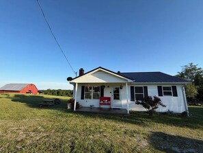 A single-story house with a front porch, a red barn in the background, and a clear sky.