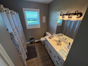 Un baño con suelo de madera, inodoro blanco, lavabo color beige con espejo y ventana con persianas.
