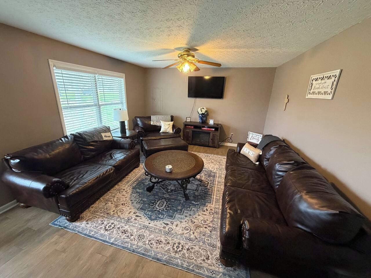 A living room with a brown leather sofa set, a coffee table, a patterned rug, a ceiling fan, and a flat-screen TV mounted on the wall.
