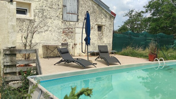 Une piscine avec des chaises longues, un bâtiment en pierre et une clôture verte.