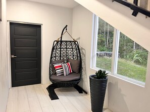 Ein Zimmer mit einer schwarzen Tür, einem Hängestuhl mit Kissen, einer Topfpflanze und einem Fenster mit Blick auf Grünflächen.