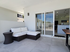 Ein moderner Außenbereich mit einer Sitzecke, inklusive Sofa und kleinem Tisch, eine Schiebetür zu einem Balkon und ein durch die Tür einsehbarer Küchenbereich.