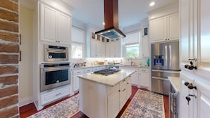 A kitchen with white cabinetry, stainless steel appliances, a central island, and a brick accent wall. - Spirited Garden- Historic/Central to everything (Galveston)