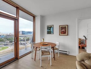 Ein Zimmer mit Esstisch und Stühlen, einer Couch, einem großen Fenster mit Stadtblick und zwei gerahmten Bildern an der Wand.