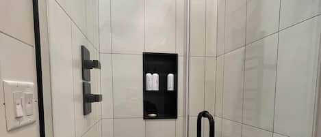 Um chuveiro moderno com porta de vidro, um banquinho de madeira e um prateleira com artigos de higiene pessoal.