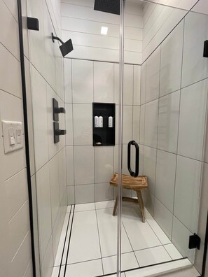 Una ducha moderna con puerta de vidrio, un taburete de madera y un estante con artículos de aseo.