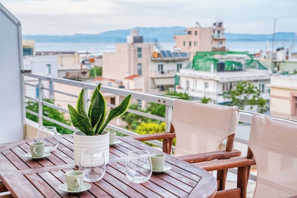A wooden table with a potted plant, two cups, and two glasses on a balcony overlooking a cityscape with buildings and a body of water. - Chios Comfort Home D1 (Chios)