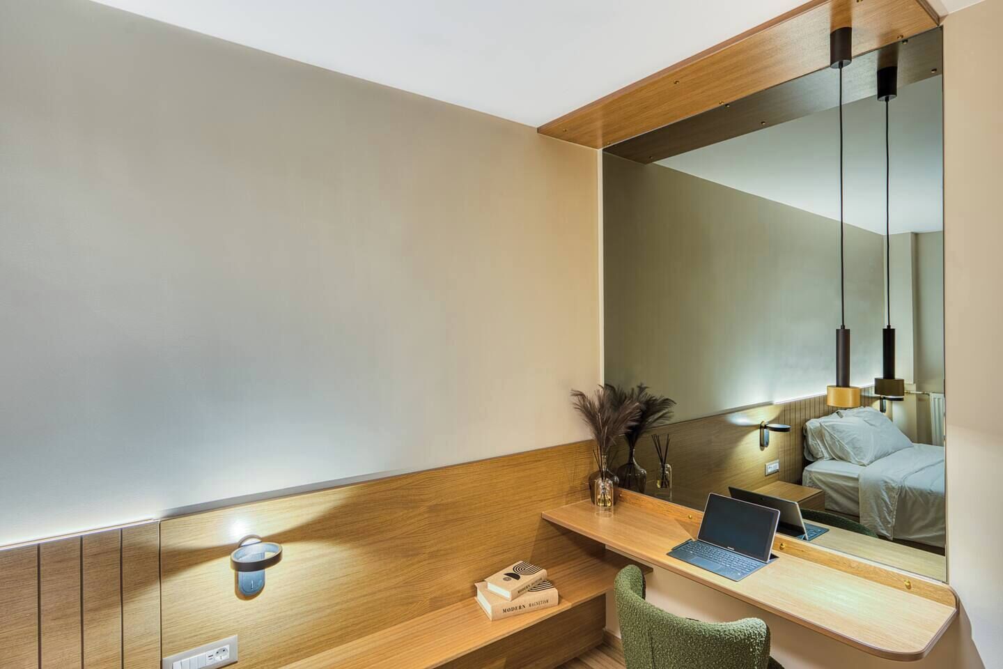 A modern hotel room with a wooden desk, a laptop, a green chair, a bedside table, and a large mirror.