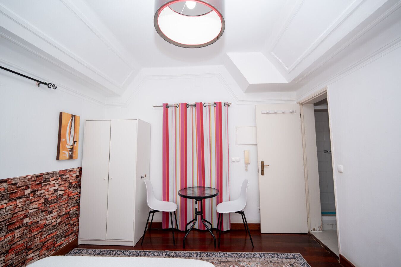 A compact room with a bed, a small table, two chairs, a striped curtain, a painting, and a brick-patterned wall.