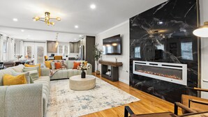 A modern living room with a sectional sofa, a fireplace, a dining area, and a kitchen in the background.
