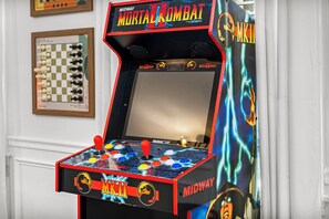Máquina de arcade de Mortal Kombat com joystick e botões, posicionada diante de uma parede branca com quadros decorativos.