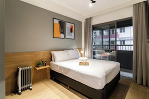 Une chambre d’hôtel moderne avec un grand lit, un radiateur, une petite table de chevet agrémentée d’une plante, et donnant sur le balcon extérieur.