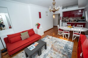 Ein Wohnzimmer mit rotem Sofa, einem Esstisch mit weißen Stühlen, einer Küche mit roten Schränken und einer Kronleuchter.