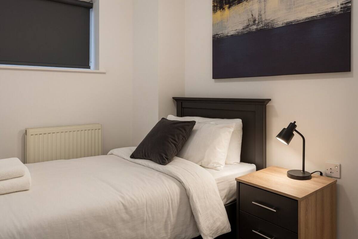 A bedroom with a bed, a bedside table with a lamp, a radiator, and a painting on the wall.