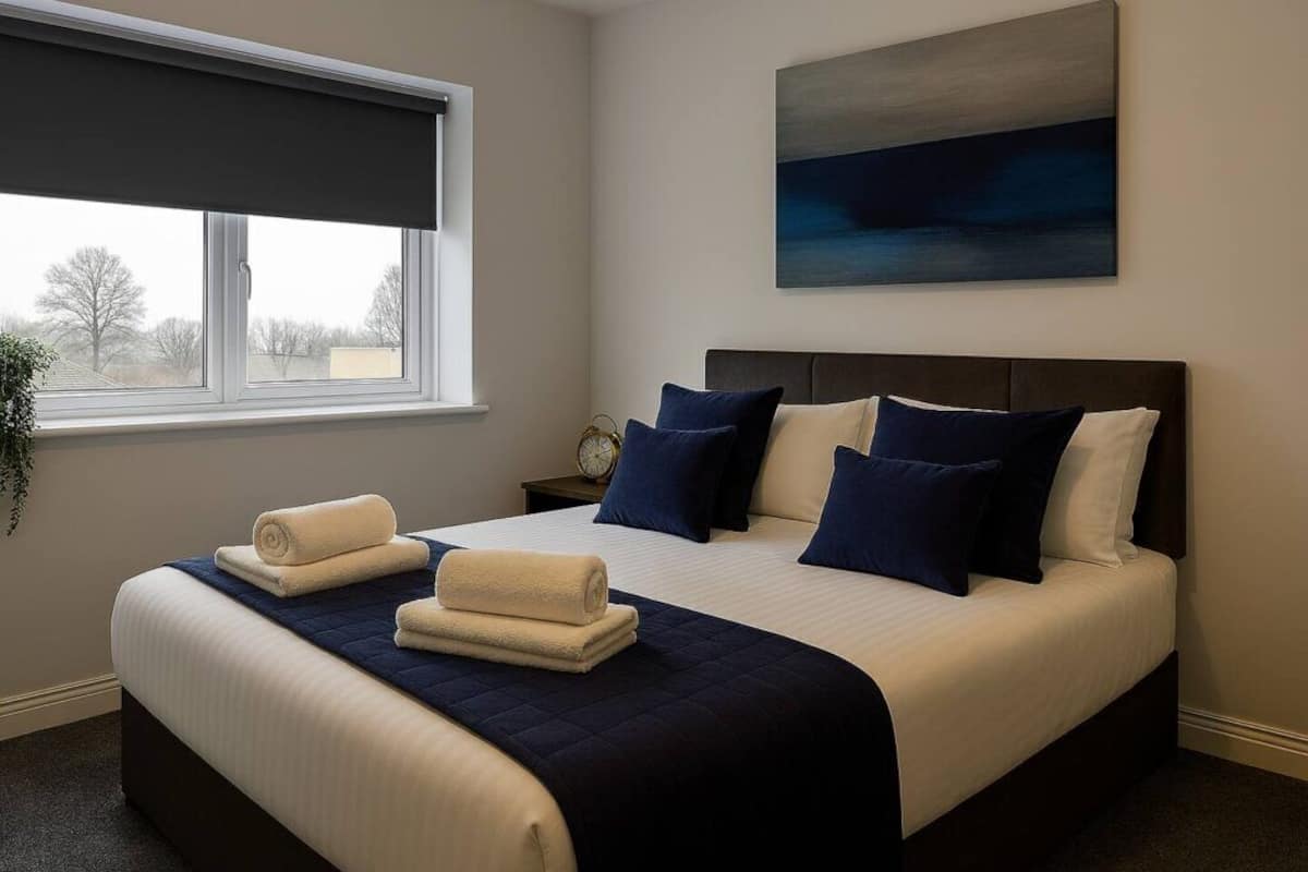 A neatly made bed with a dark blue bedspread and pillows, a window with a dark blind, and a painting on the wall.