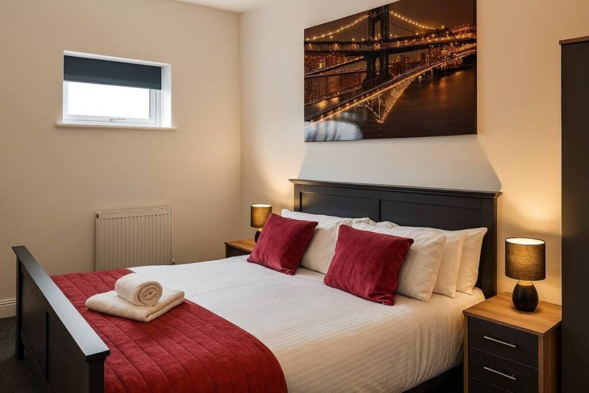 A hotel room with a bed, a window, a nightstand, a lamp, and a painting of a bridge.