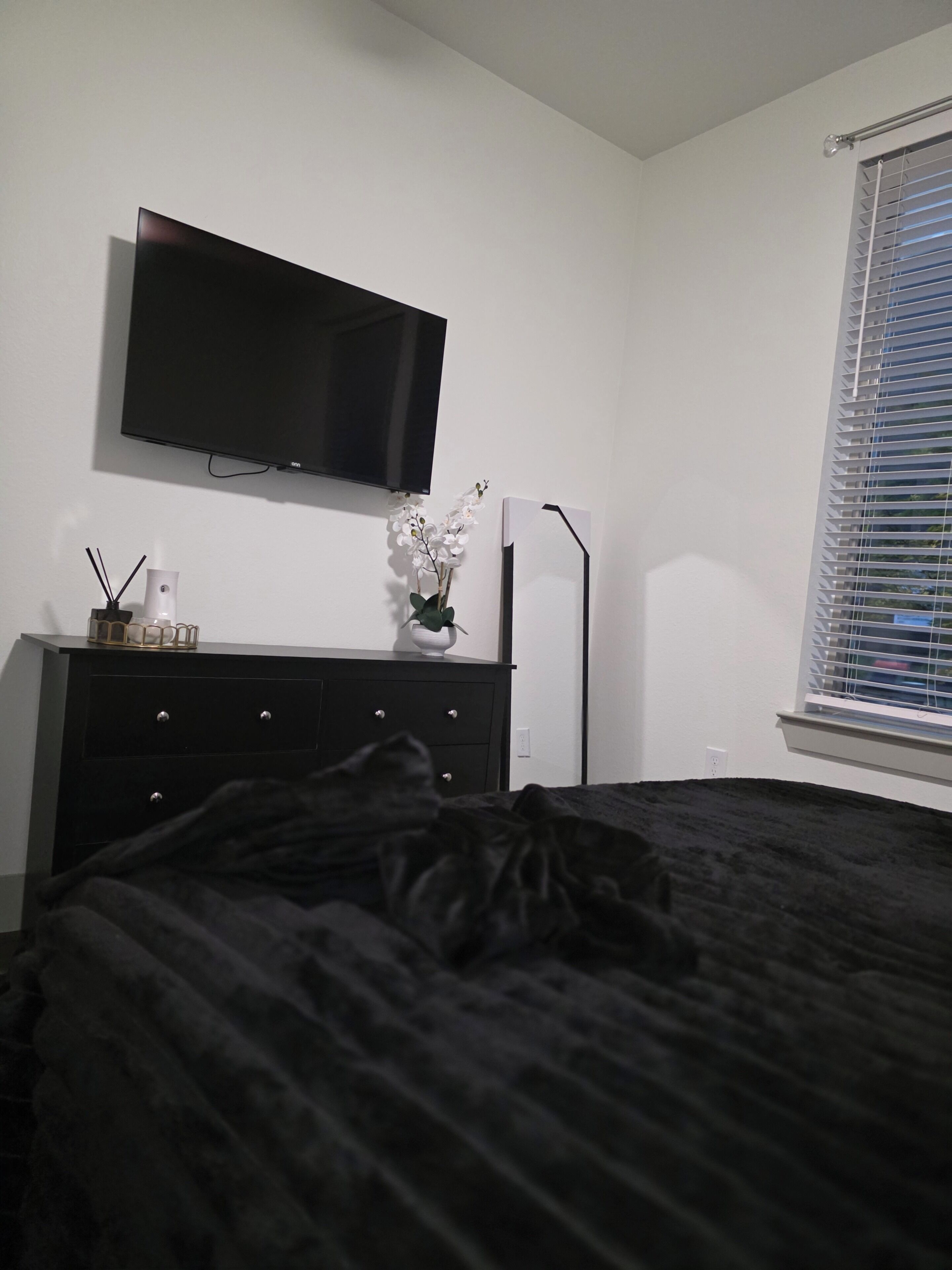 A bedroom with a bed, a dresser, a TV mounted on the wall, a window with blinds, and a floor lamp.