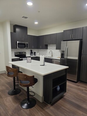 A modern kitchen with dark cabinetry, a white island, stainless steel appliances, and two bar stools. - Heart of Addison
In a cozy, friendly and safe community. (Addison)