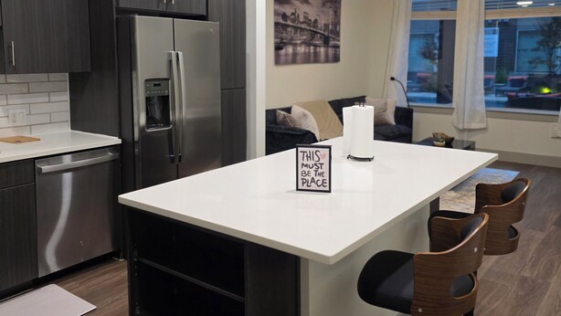 A modern kitchen with dark cabinets, a stainless steel refrigerator, a white island with bar stools, and a cityscape painting on the wall. - Heart of Addison
In a cozy, friendly and safe community. (Addison)