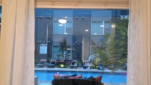 A view through a window with blinds, a pool, outdoor seating, and a building with multiple windows and lights. - Heart of Addison
In a cozy, friendly and safe community. (Addison)