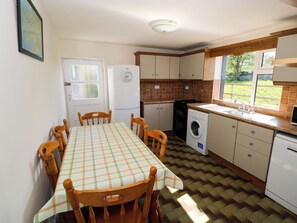 A compact kitchen with a dining table, chairs, a refrigerator, a washing machine, and a window with a view of greenery. - Cahir Cottage (Ballinrobe)