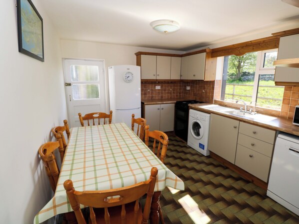 A compact kitchen with a dining table, chairs, a refrigerator, a washing machine, and a window with a view of greenery. - Cahir Cottage (Ballinrobe)