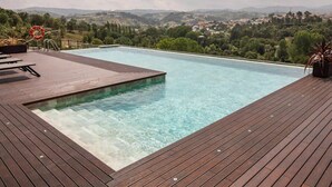 Une piscine rectangulaire à l’eau claire, entourée d’une terrasse en bois, et offrant une vue panoramique sur un paysage pittoresque avec des collines et des bâtiments.