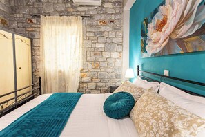 A bedroom with a stone wall, a bed with patterned bedding, a teal throw blanket, and a floral painting on the wall. - Beachfront Mani Luxury Stone Nest, Steps from Sea (Kokkala)