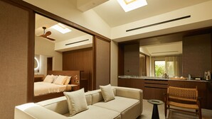 A modern hotel room with a sofa, a small round table, a wicker chair, a kitchen area, and a bed with a headboard.