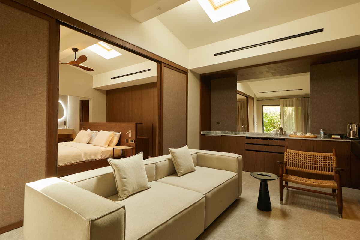 Una habitación de hotel moderna con sofá, una pequeña mesa redonda, una silla de mimbre, una zona de cocina y una cama con cabecera.