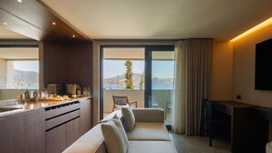 A modern living room with a sofa, a dining area, and a kitchen. There is a large window offering a view of the sea and hills.