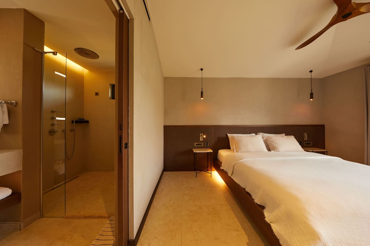 Habitación de hotel con ducha envuelta en vidrio, cama con ropa blanca, mesita de noche y ventilador de techo.