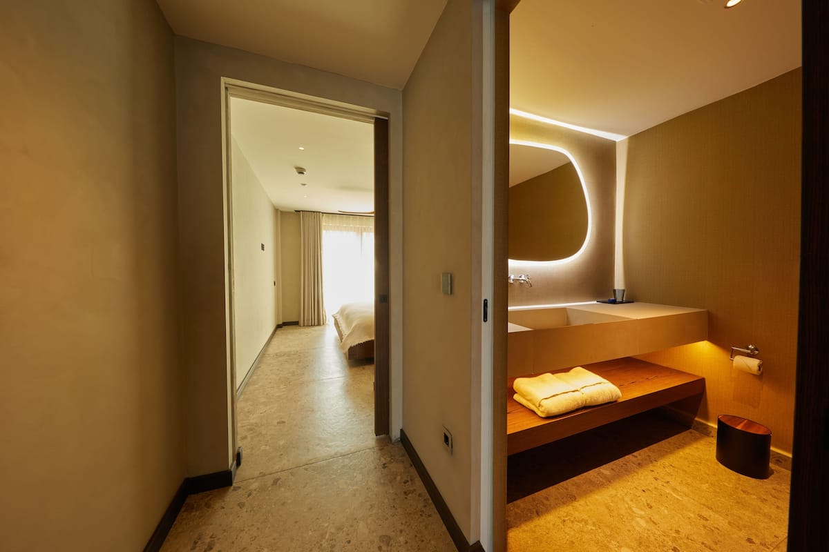 Una habitación de hotel moderna con una cama grande, un baño con un espejo amplio y un banco de madera.