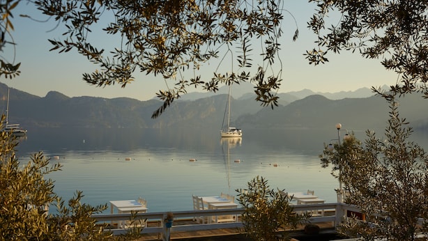 A waterfront view with a boat, tables, and chairs, surrounded by mountains.